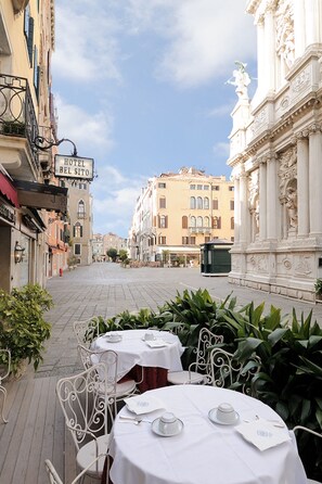 Un patio con mesas y sillas, un cartel del Hotel Bel Sito y arquitectura clásica.