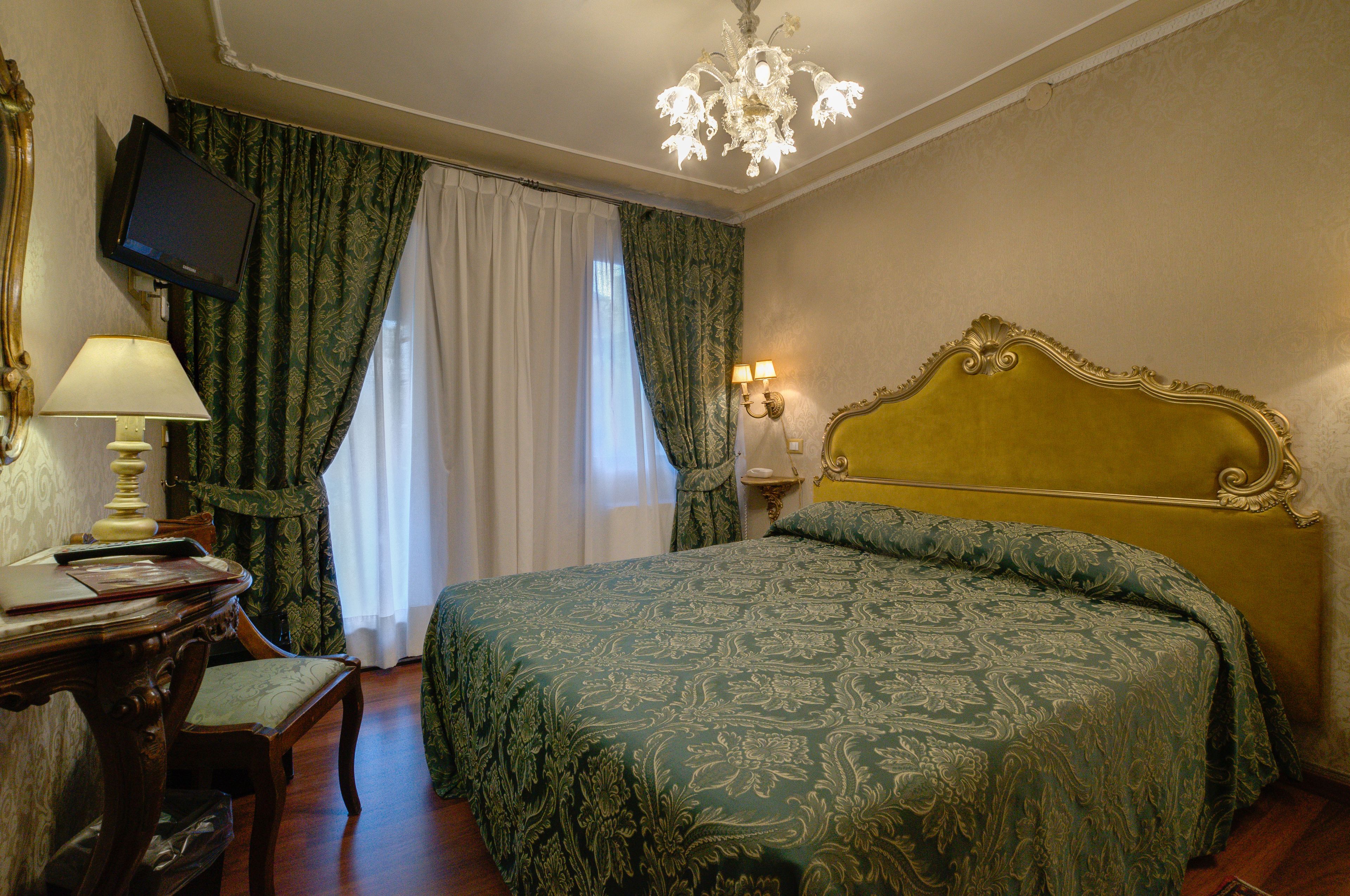 A room with a large bed, a desk with a lamp, a television on the wall, and a chandelier.