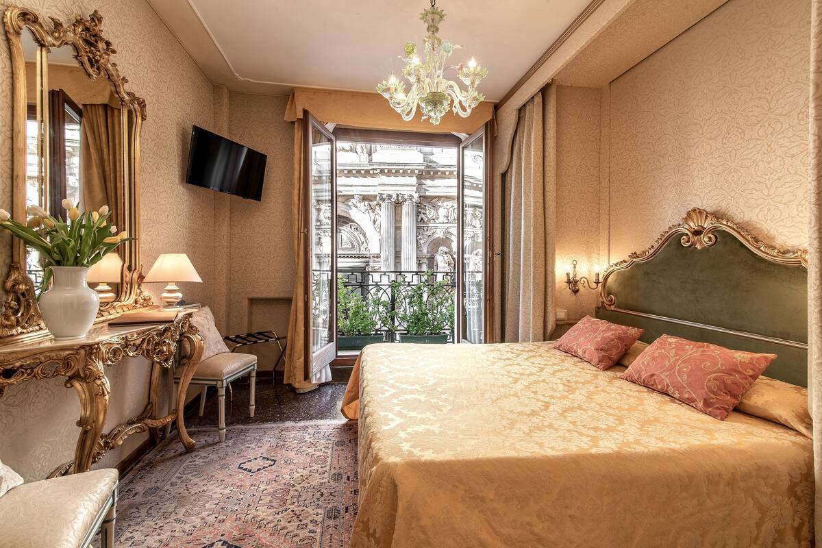 A luxurious hotel room with a large bed, ornate furniture, a chandelier, and a view of an outdoor balcony.