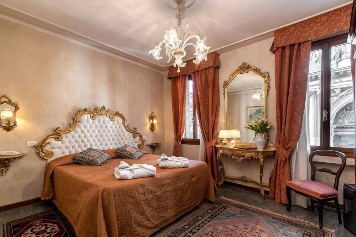 A hotel room with a large bed, a chandelier, a mirror, a desk, and a chair.