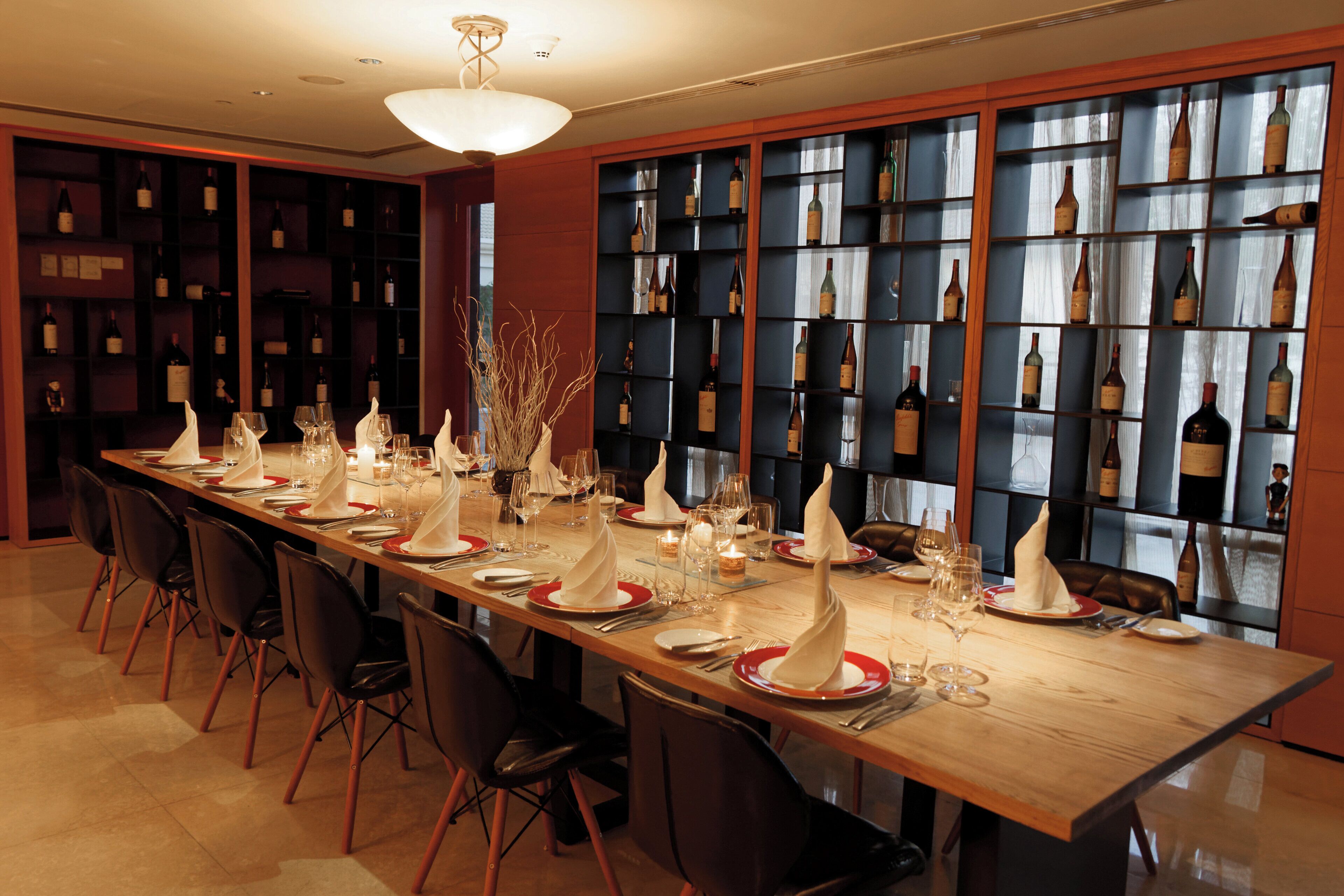 A long dining table set for a meal with chairs, wine bottles on shelves, and a modern chandelier.