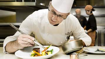 Un chef, vêtu d'un uniforme blanc et coiffé d'un haut-de-forme, prépare un plat sur une assiette blanche, agrémenté de légumes et de viande.