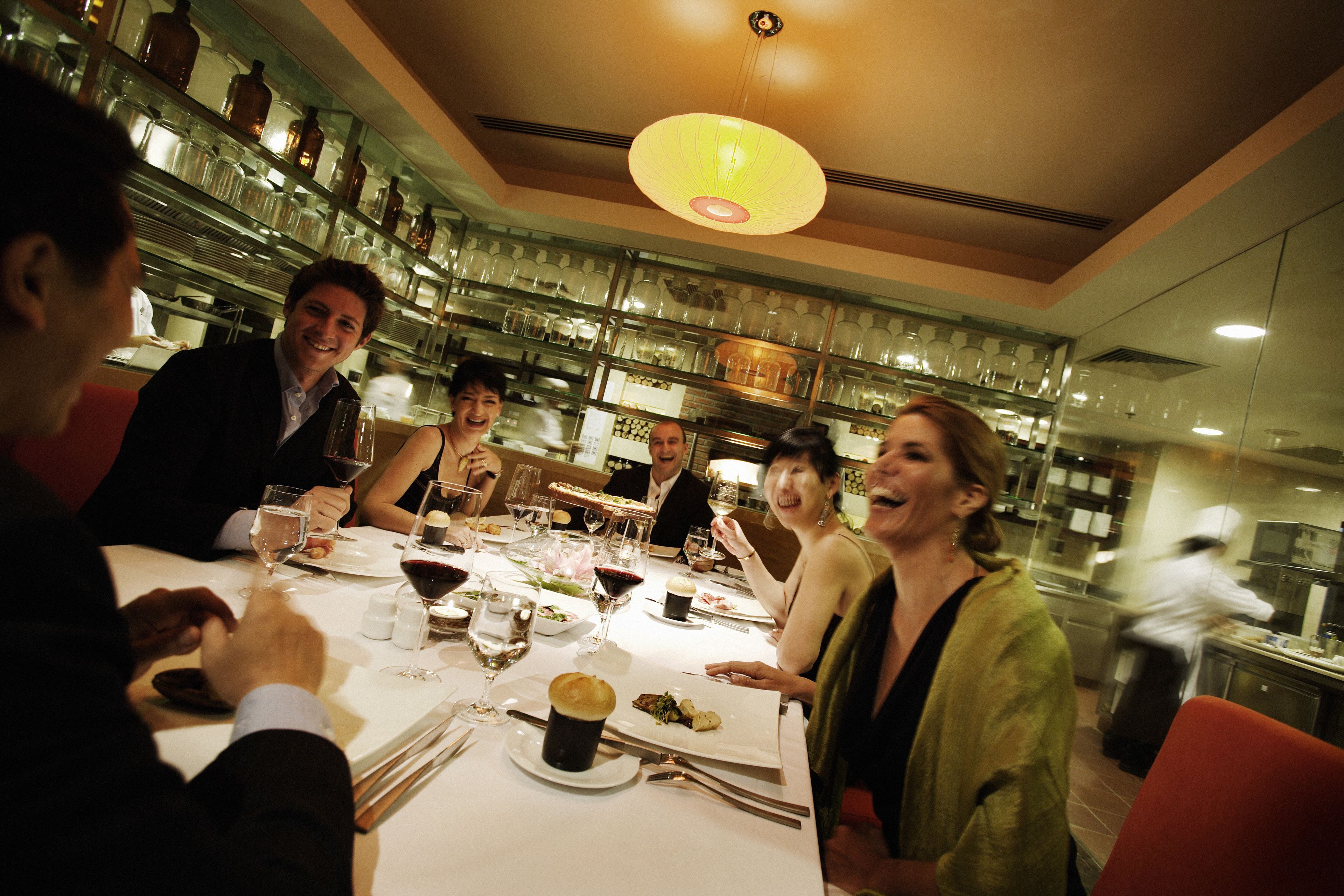 Un grupo de personas cenando en un restaurante con una decoración moderna, que incluye una zona de bar con estantes llenos de botellas y vasos.
