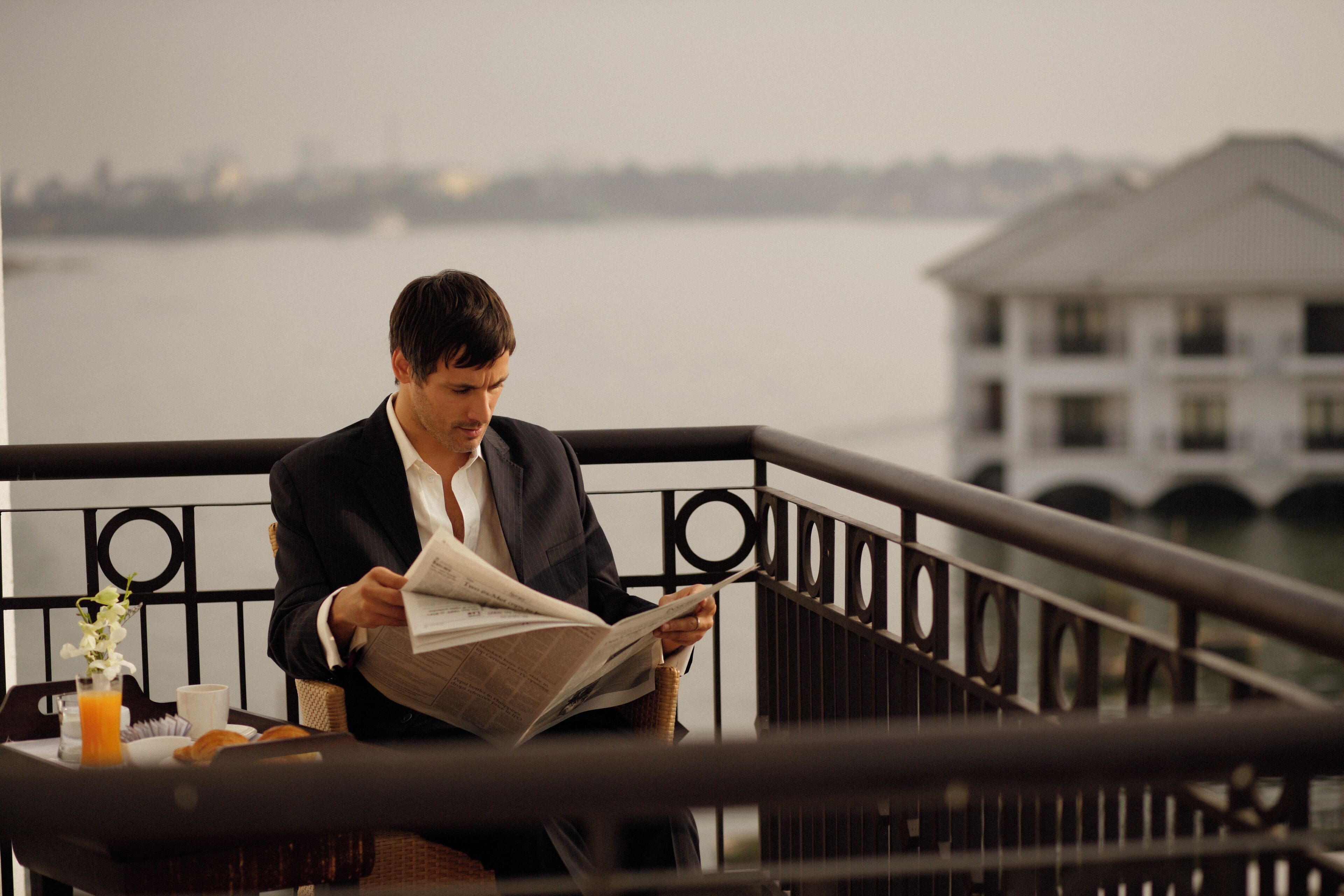 Un hombre leyendo un periódico en un balcón con barandilla, con vista a un cuerpo de agua y un edificio al fondo.