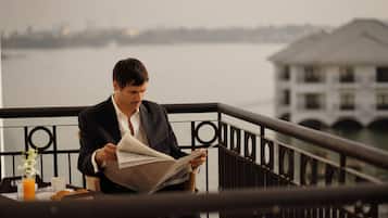 Un hombre leyendo un periódico en un balcón con barandilla, con vista a un cuerpo de agua y un edificio al fondo.
