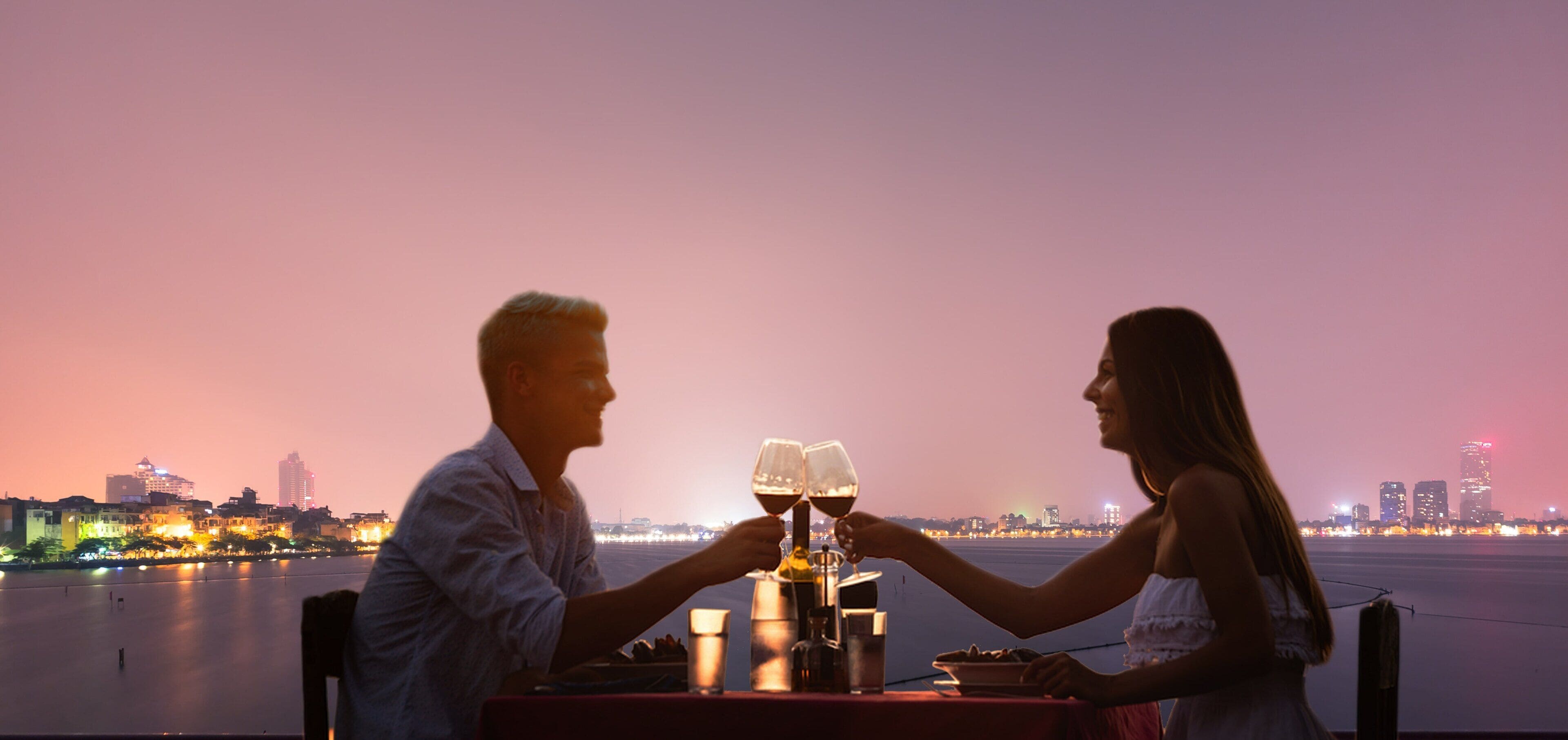 A couple toasting with wine glasses at a table with a cityscape view in the background.