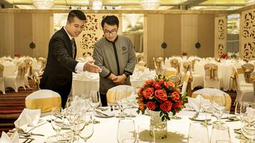 A banquet hall with round tables set for a formal event, featuring white tablecloths, gold sashes, and a centerpiece of red roses.