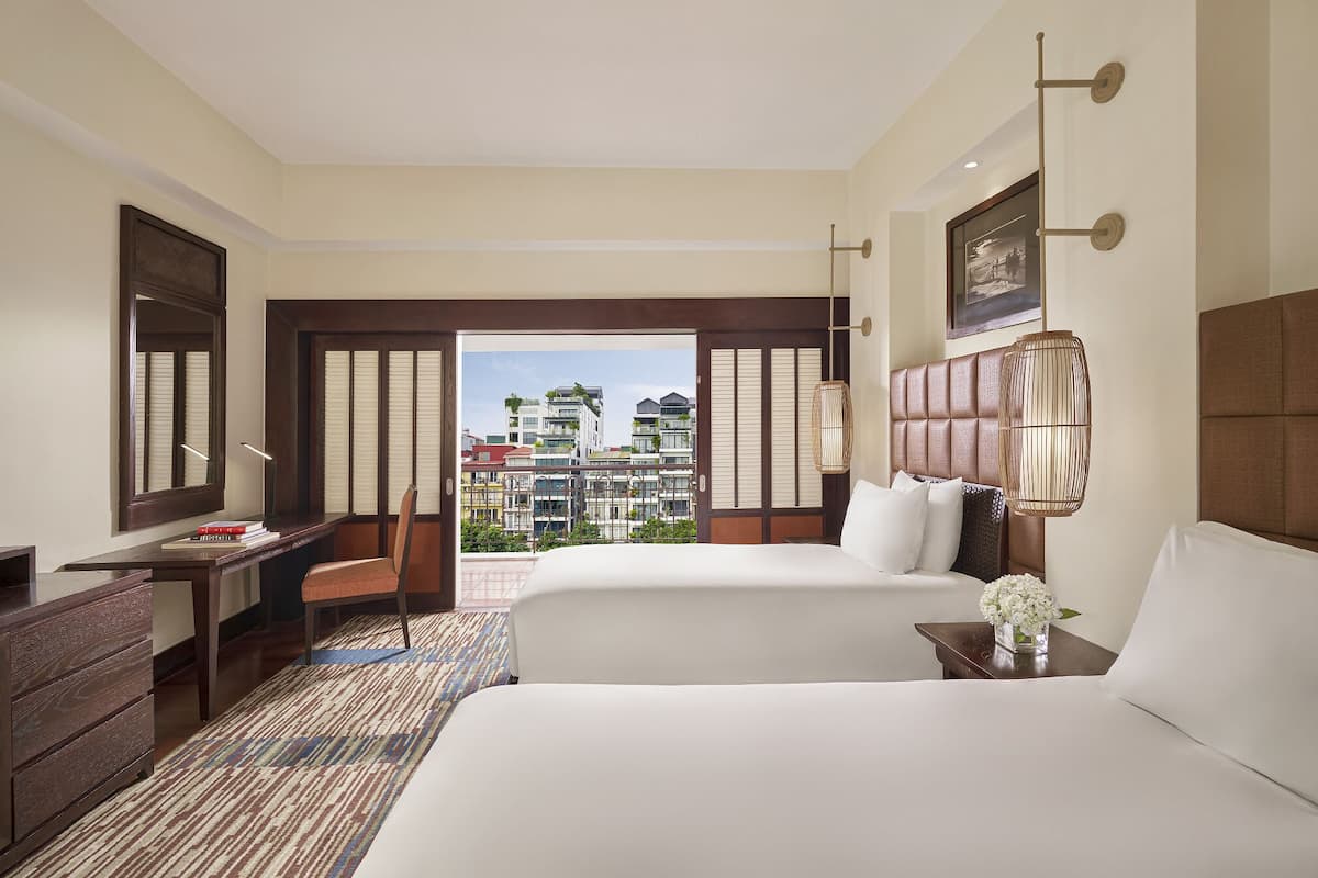 A hotel room with two beds, a desk, a chair, a large window with a view of buildings, and a hanging light fixture.