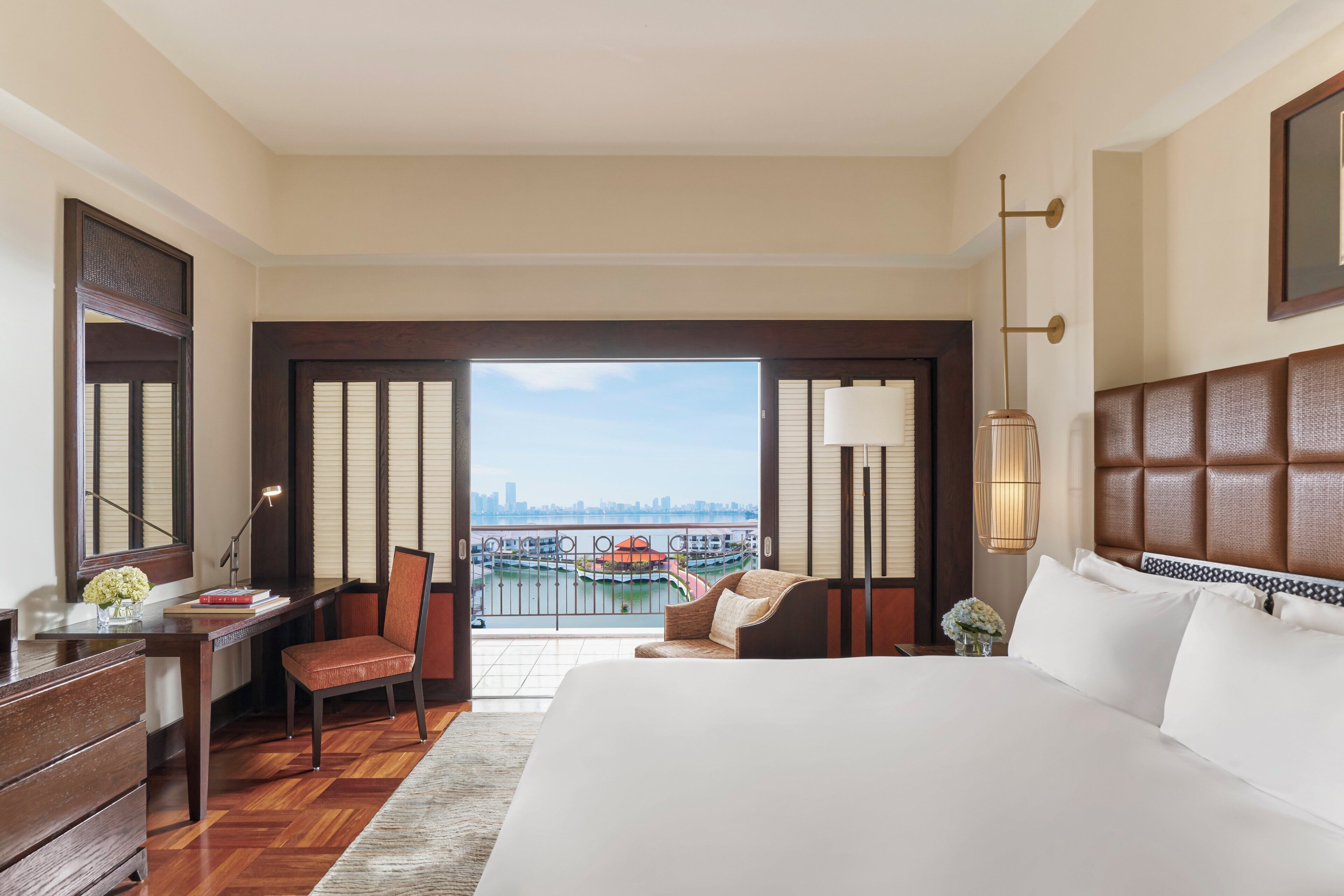 A hotel room with a large bed, a desk, a chair, a view of a cityscape and a body of water.