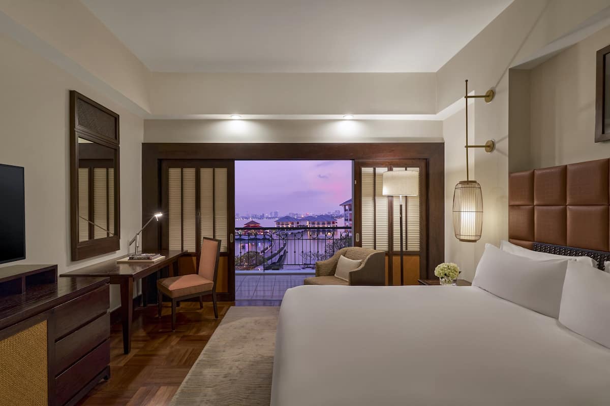 A hotel room with a bed, desk, chair, and a view of the cityscape through the balcony doors.