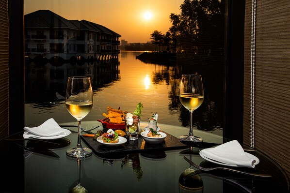 Een tafel met twee glazen witte wijn, een saladekom en een klein bord eten, met uitzicht op een rivier bij zonsondergang.