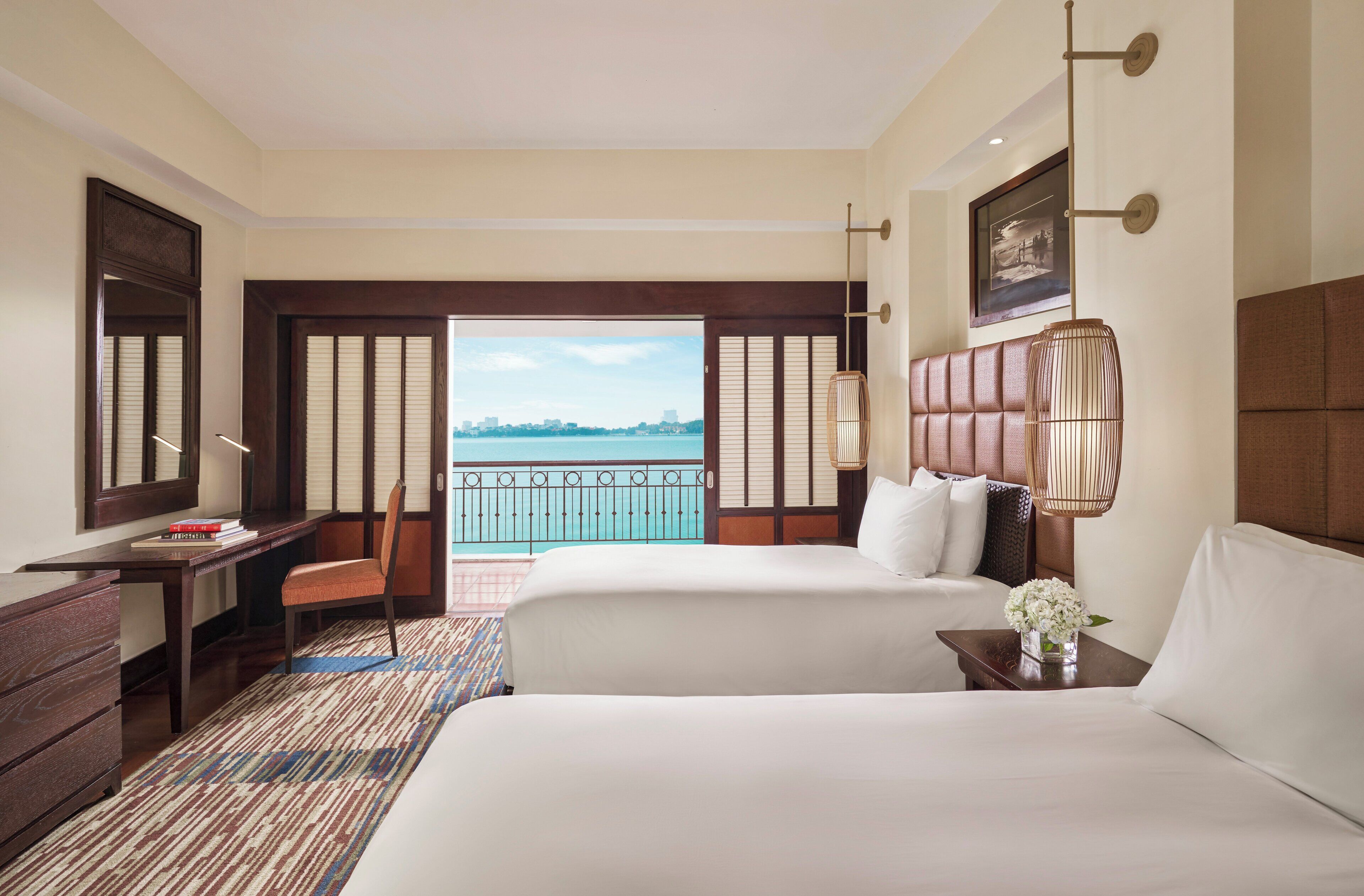 A hotel room with two beds, a desk, a chair, and a view of the sea.
