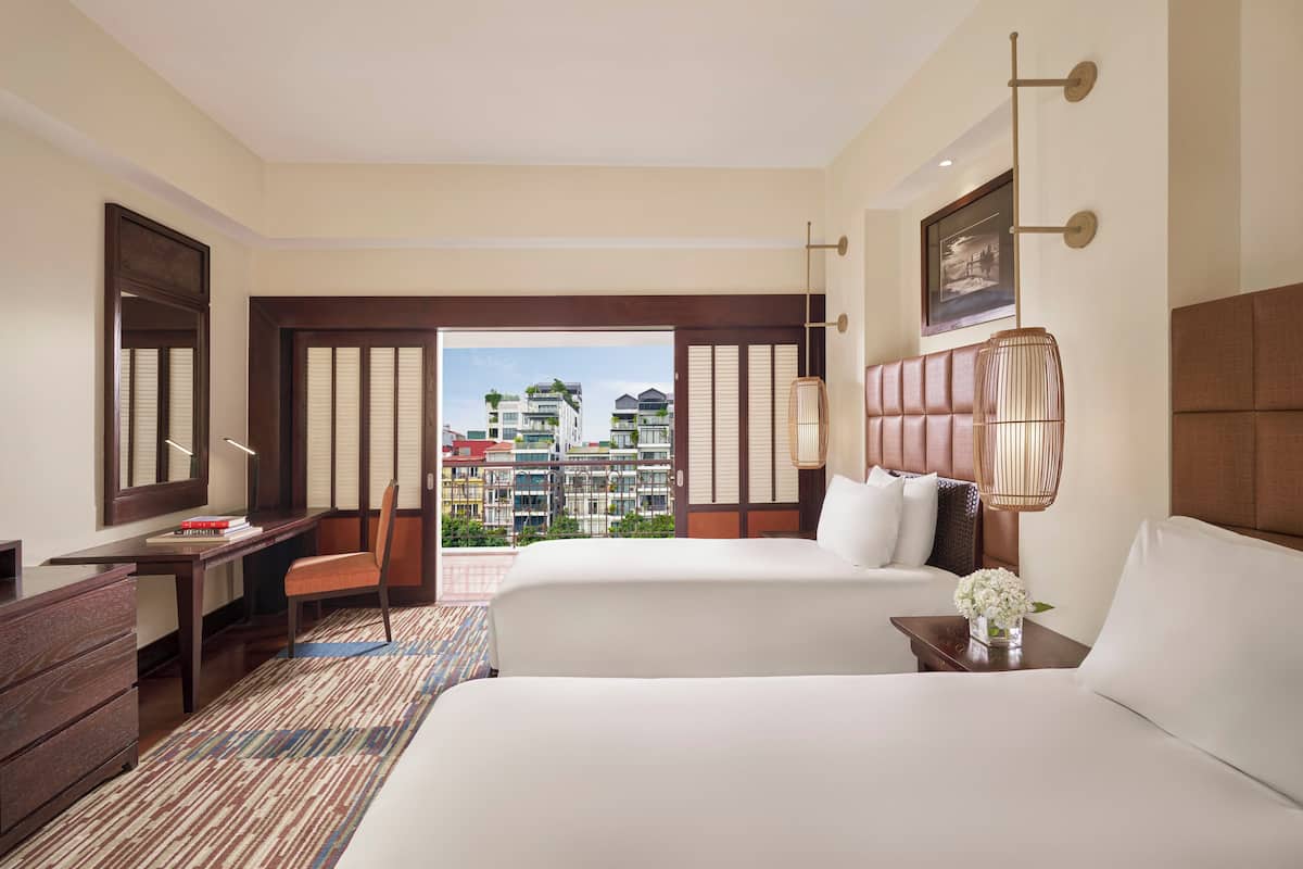 A hotel room with two beds, a desk, a chair, a lamp, and a view of buildings.