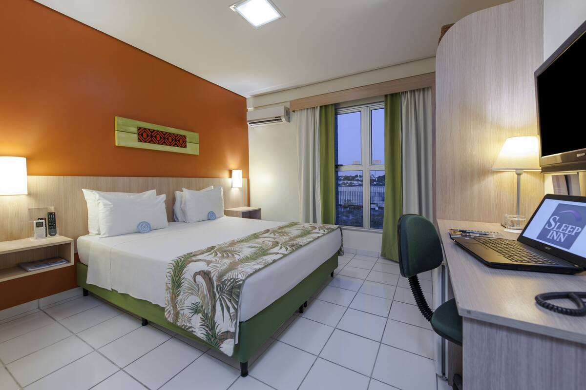 A hotel room with a bed, a desk with a laptop, a TV, and a view of the city through the window.