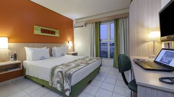 A hotel room with a bed, a desk with a laptop, a TV, and a view of the city through the window.