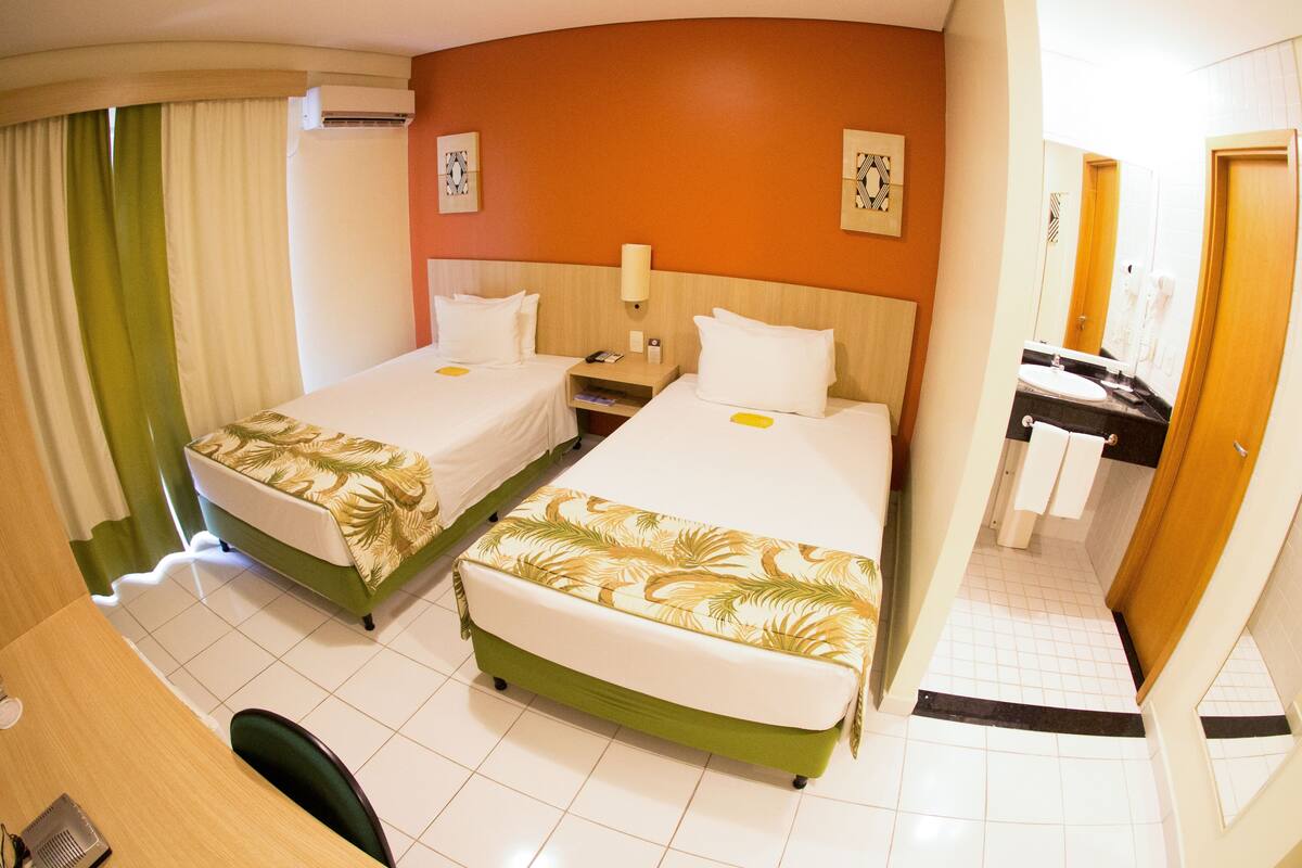 A hotel room with two beds, a desk, a chair, a bathroom with a sink and mirror, and a shower.