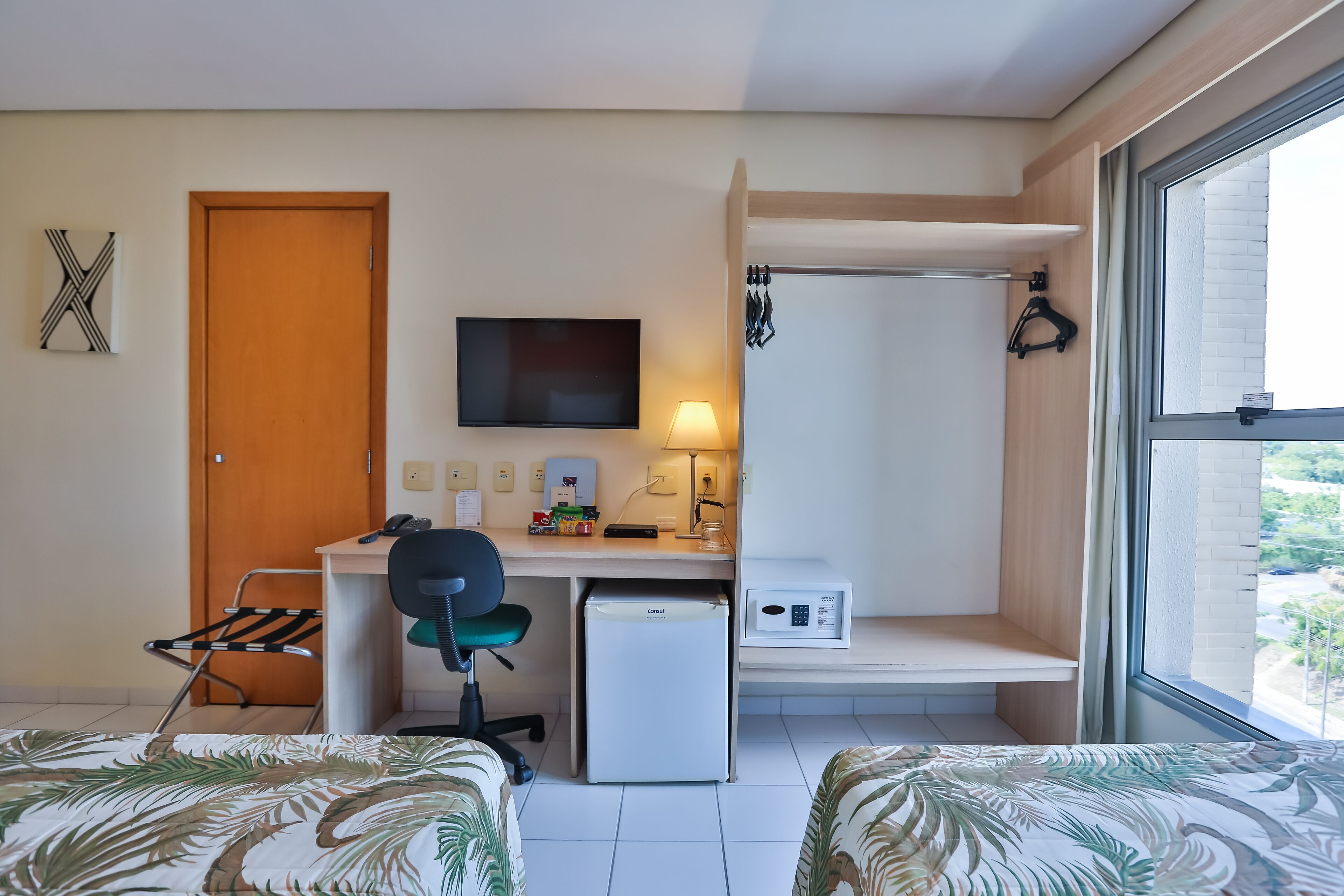 A compact hotel room with a bed, a desk with a computer, a refrigerator, and a TV.
