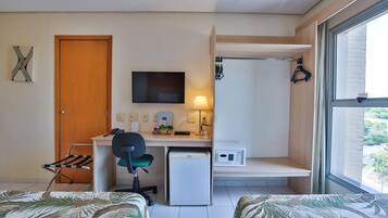A compact hotel room with a bed, a desk with a computer, a refrigerator, and a TV.