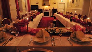 Uma sala de jantar formal com mesas longas preparadas para uma refeição, velas e lareira.