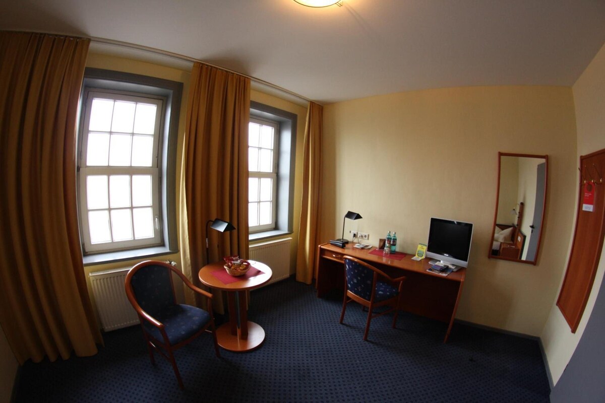 A hotel room with a desk, two chairs, a television, and a window with curtains.