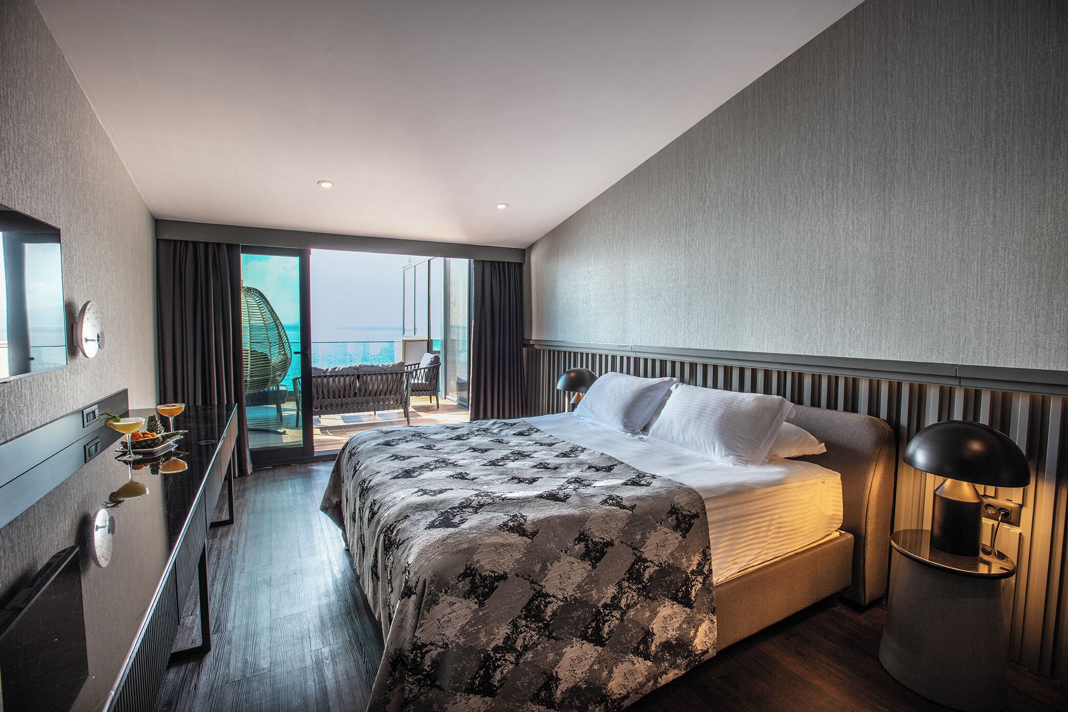 A modern hotel room with a large bed, a desk, and a view of the sea.