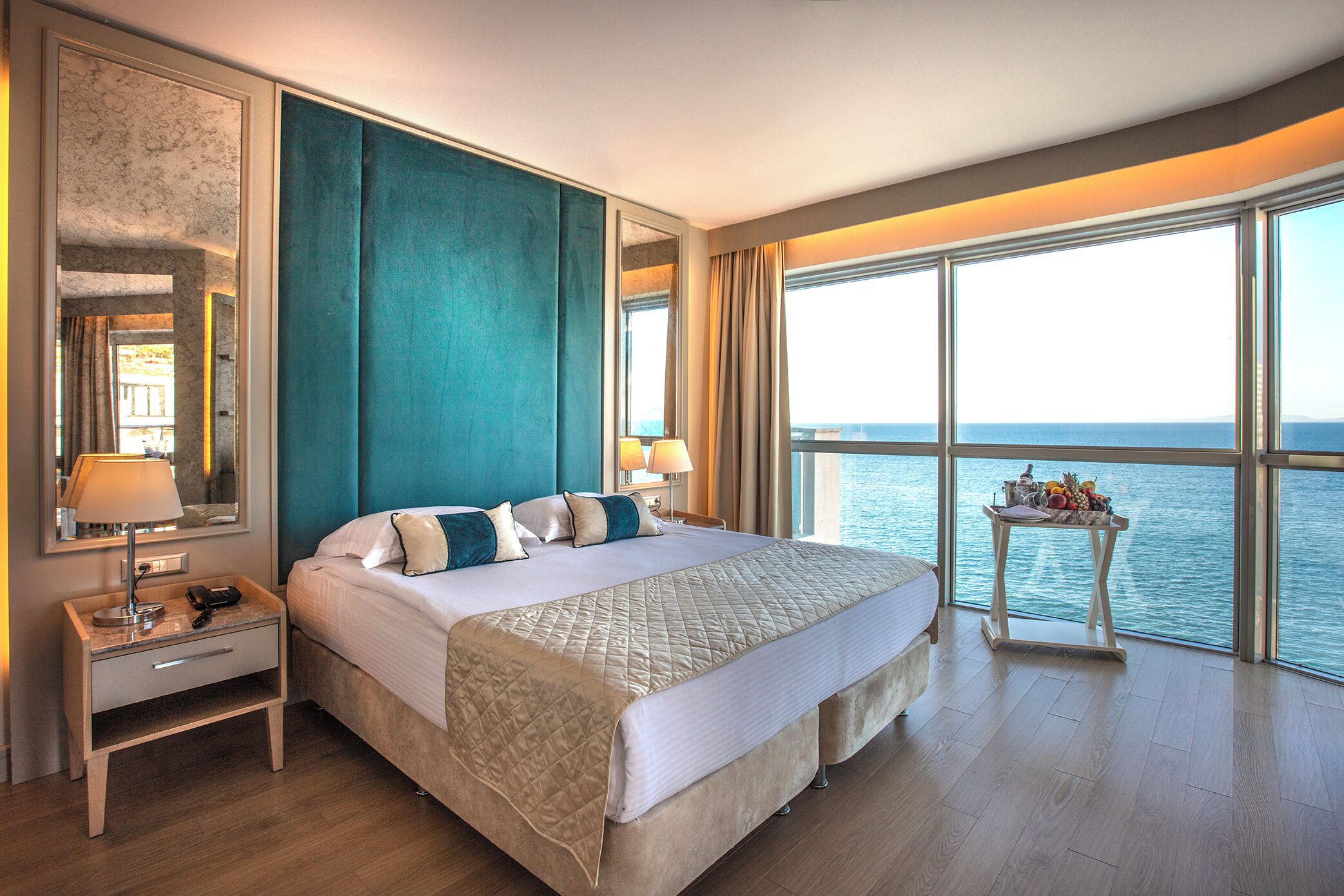 Um quarto espaçoso com uma cama grande, mesa de cabeceira, espelho e vista para o mar através de amplas janelas.