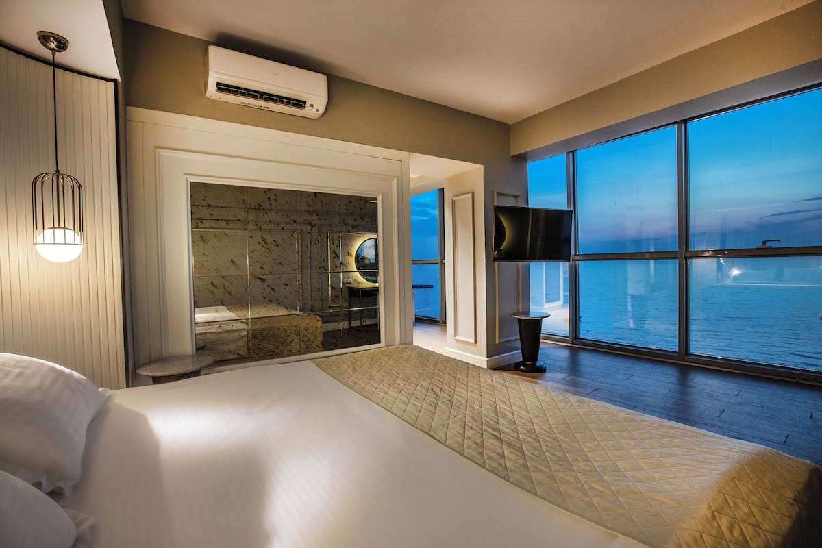 Habitación de hotel con una cama grande, vistas al mar, baño con pared de mármol y televisor de pared.
