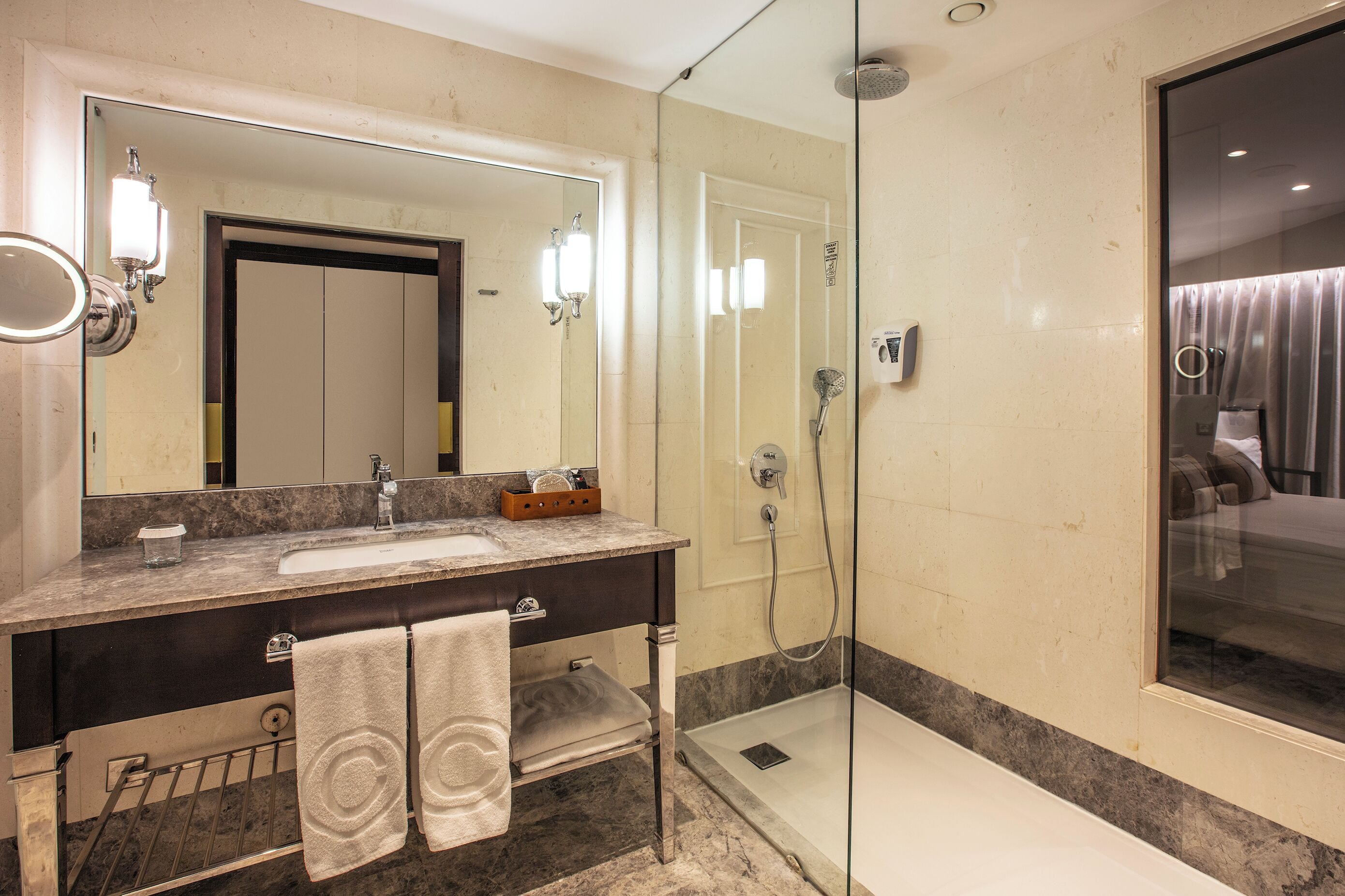 A modern bathroom with a marble sink, a large mirror, a glass-enclosed shower, and a visible door leading to a bedroom.