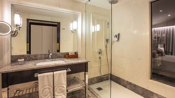 A modern bathroom with a marble sink, a large mirror, a glass-enclosed shower, and a visible door leading to a bedroom.