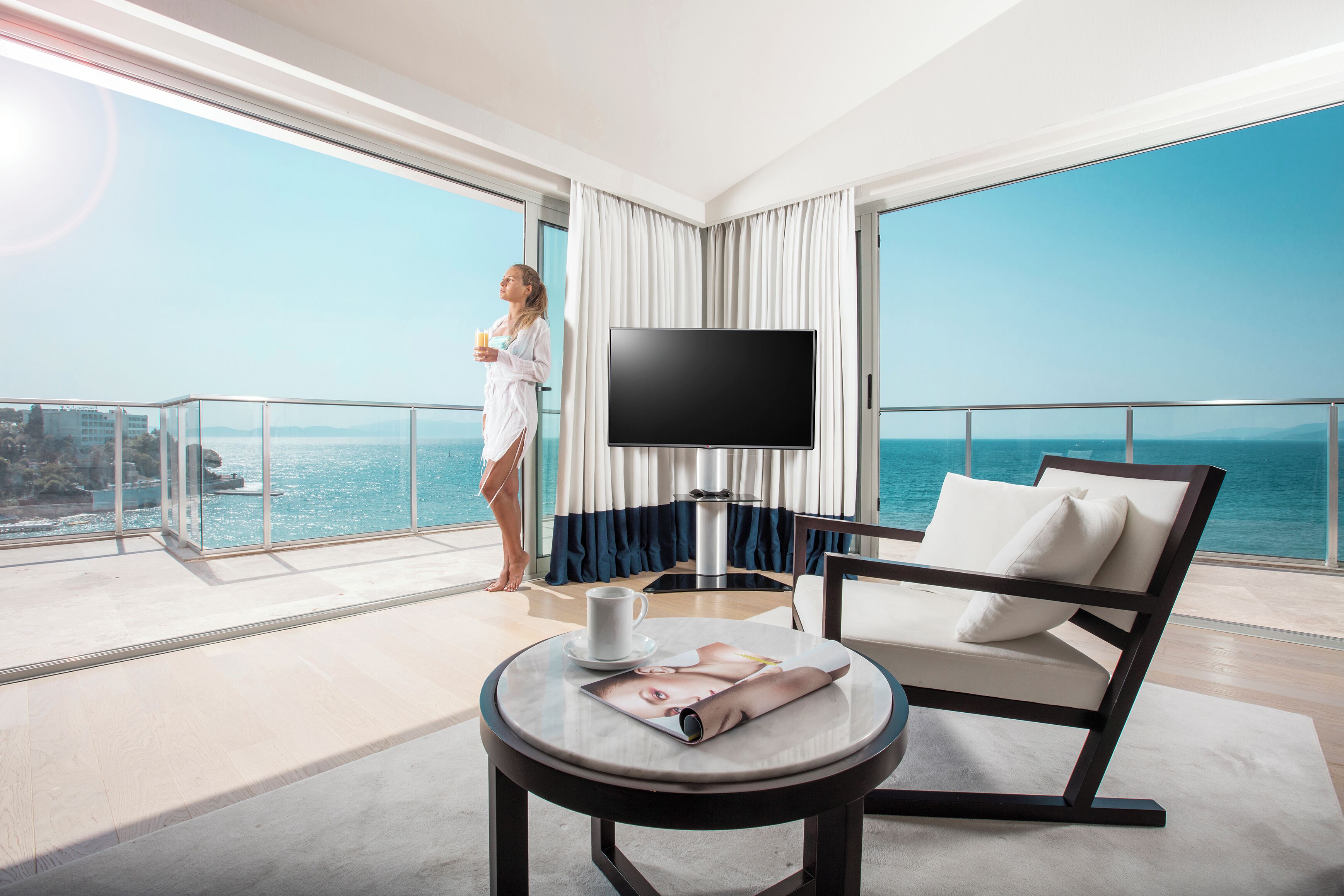 A modern hotel room with a large balcony, a flat-screen TV, a comfortable chair, and a table with a magazine.