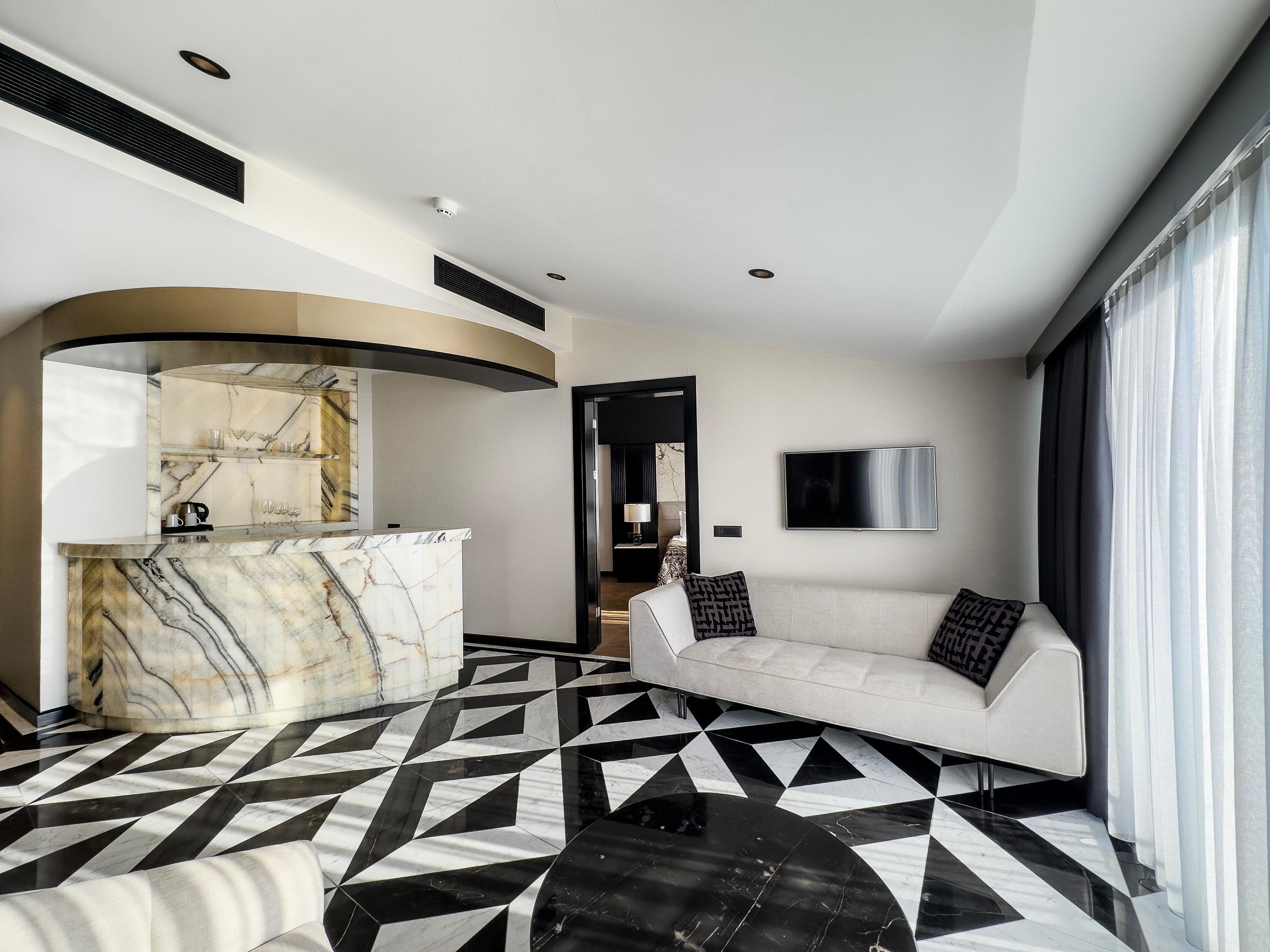 A modern hotel room with a marble reception desk, a black and white geometric floor, a white sofa with patterned cushions, and a large window with curtains.