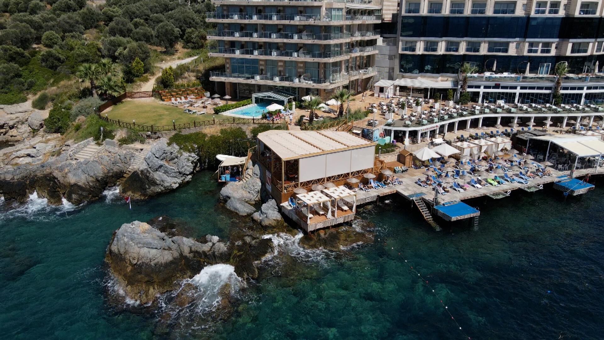 Vista aérea de un hotel costero con piscina, zona de estar exterior y un muelle que se adentra en el agua.