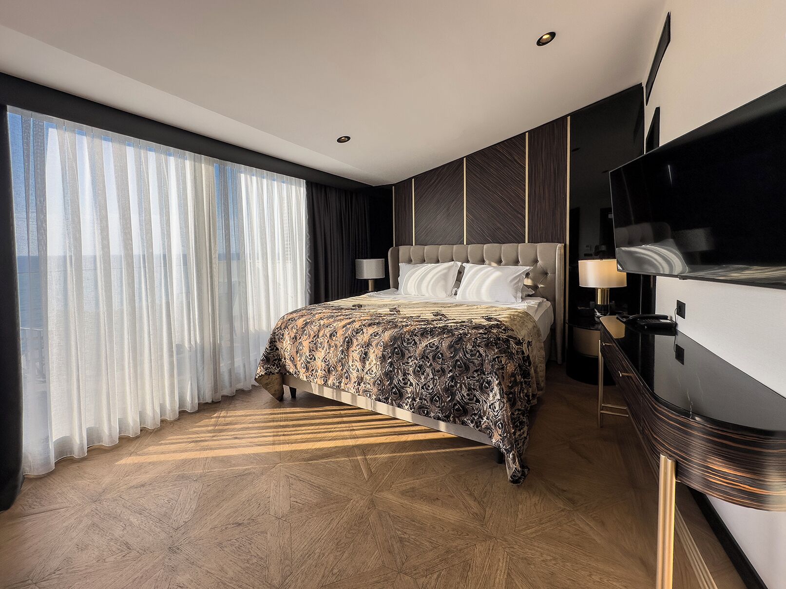 A modern bedroom with a large bed, a TV, and a view of the city through sheer curtains.