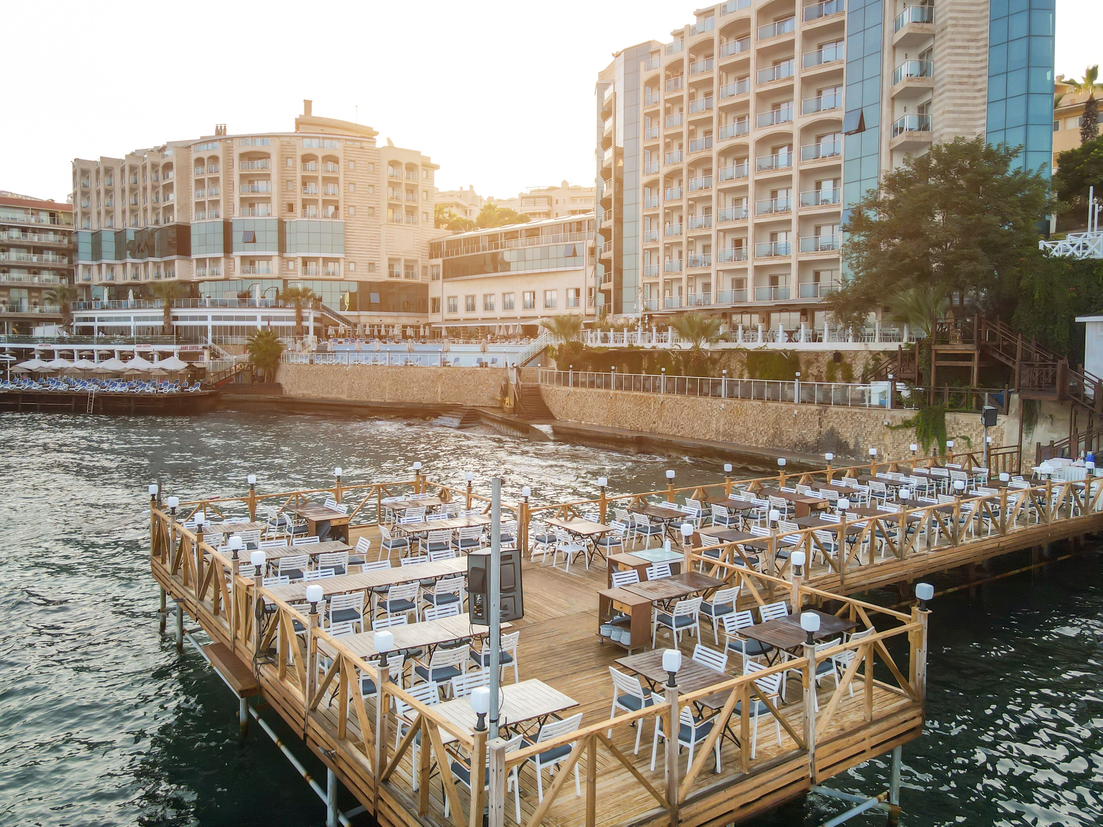 A waterfront dining area with tables and chairs on a wooden deck, overlooking a body of water.
