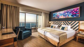 A hotel room with a large bed, a desk, a chair, a view of the sea, and a colorful painting on the wall.