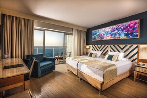 Camera d'albergo con un letto grande, una scrivania, una sedia, vista sul mare e un quadro colorato appeso al muro.
