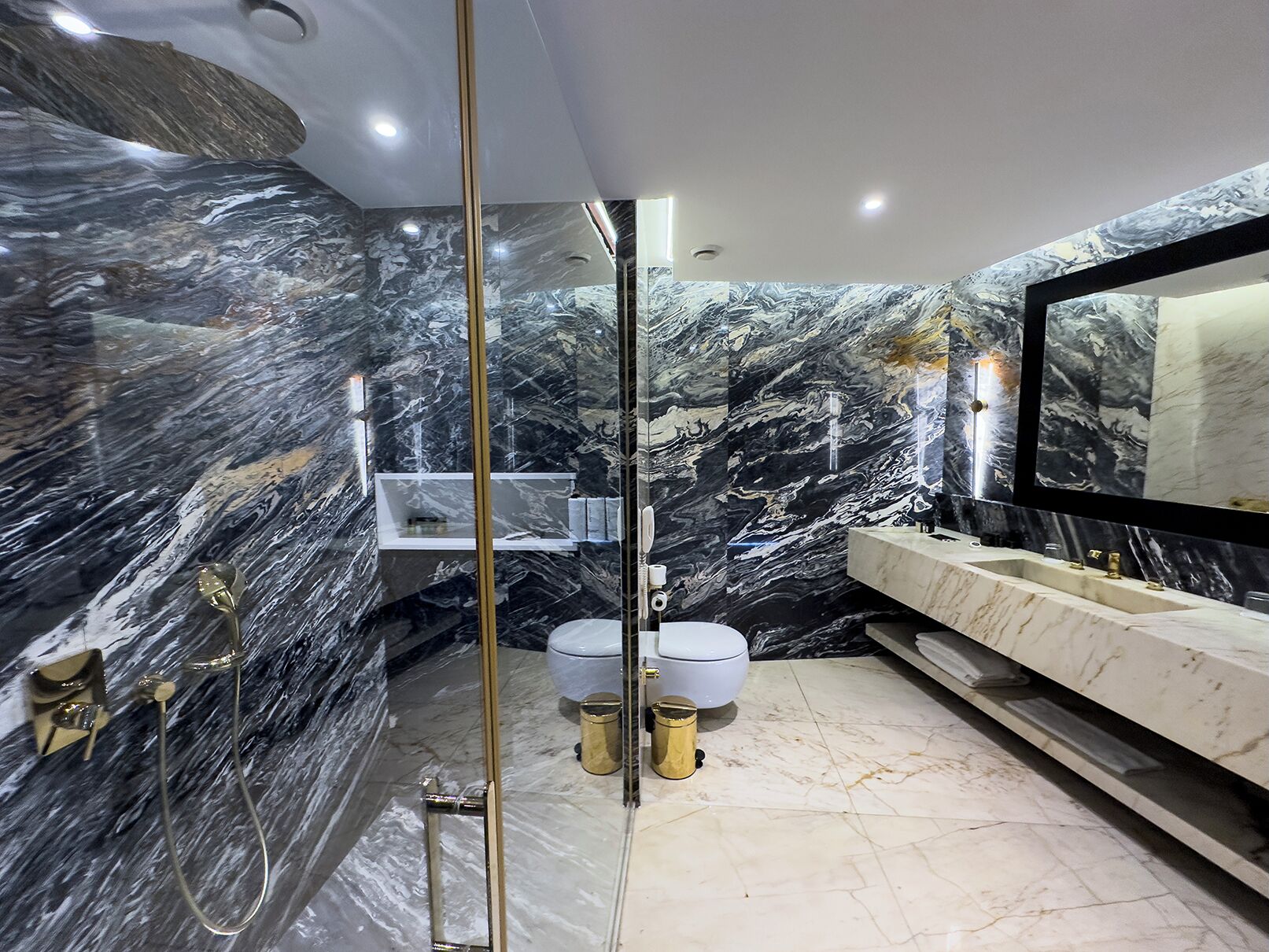 Une salle de bain moderne avec des murs en marbre, une douche en verre, des toilettes blanches et un meuble-lavabo doté d’un grand miroir.