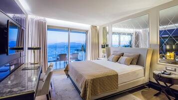 A hotel room with a large bed, a desk with a chair, a TV, and a balcony with a view of the sea.