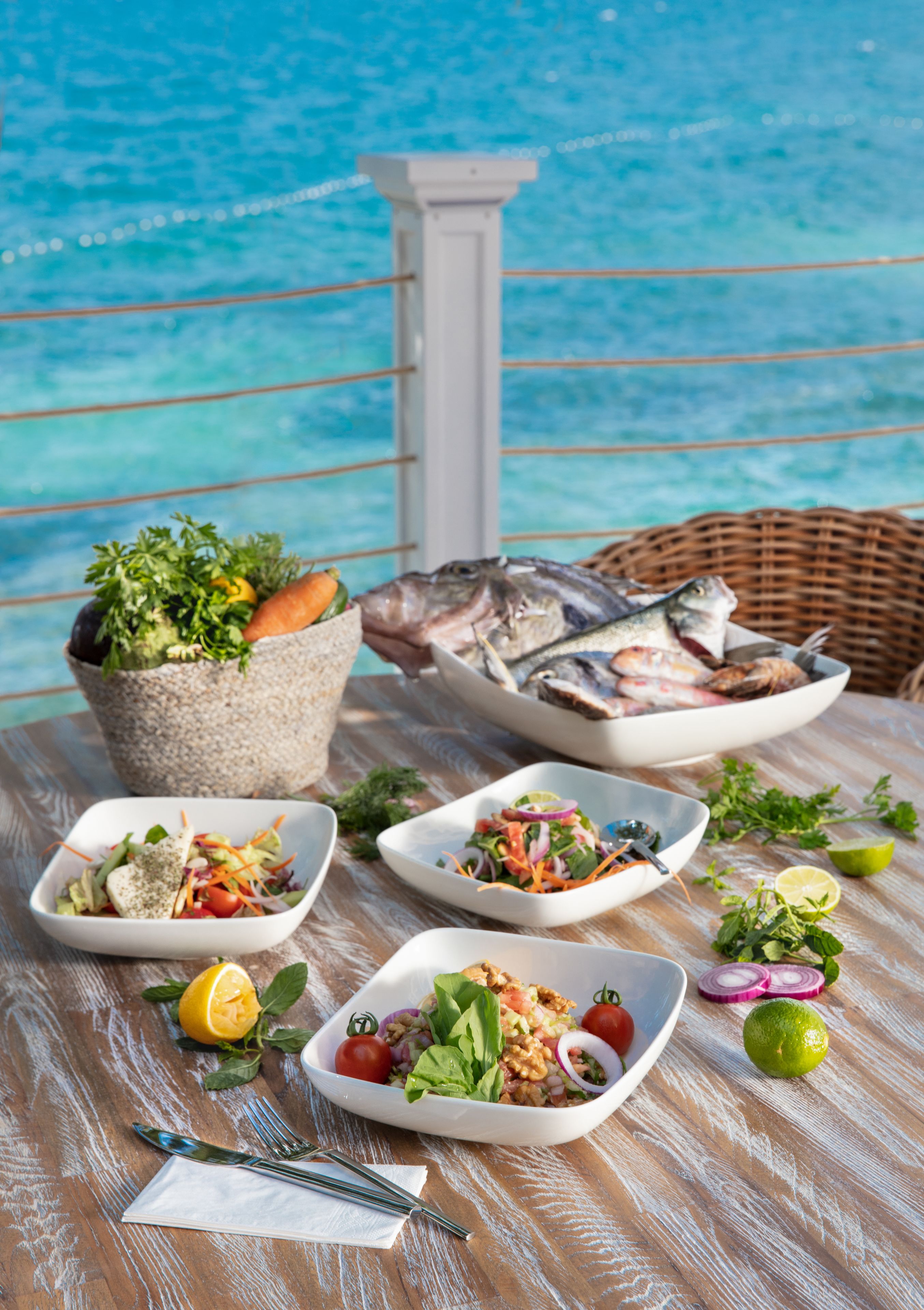 A table with various dishes and a wicker chair, set against a backdrop of a blue ocean view.