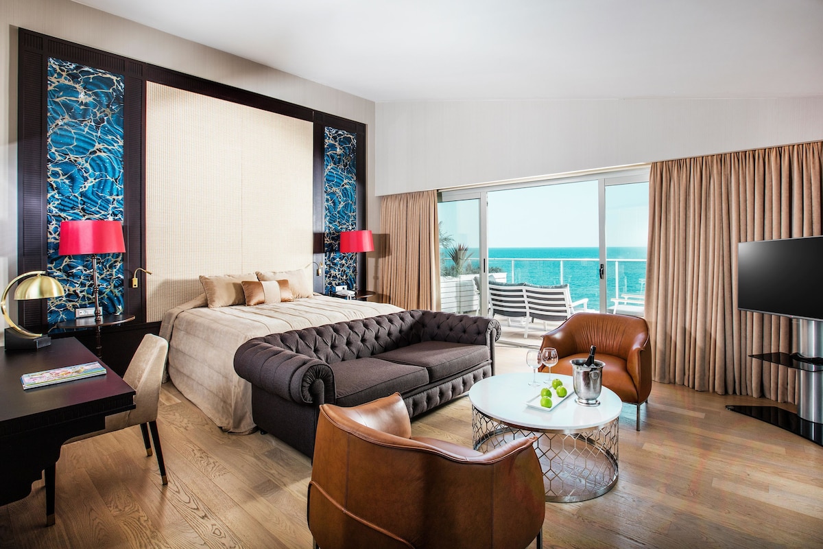 A modern hotel room with a large bed, a sofa, a desk, a TV, and a balcony with a view of the sea.