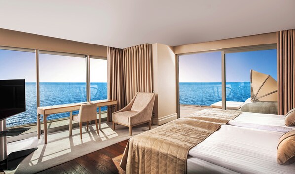 Habitación de hotel con un ventanal grande que ofrece vistas al mar, un escritorio con una silla, una cama y un televisor.