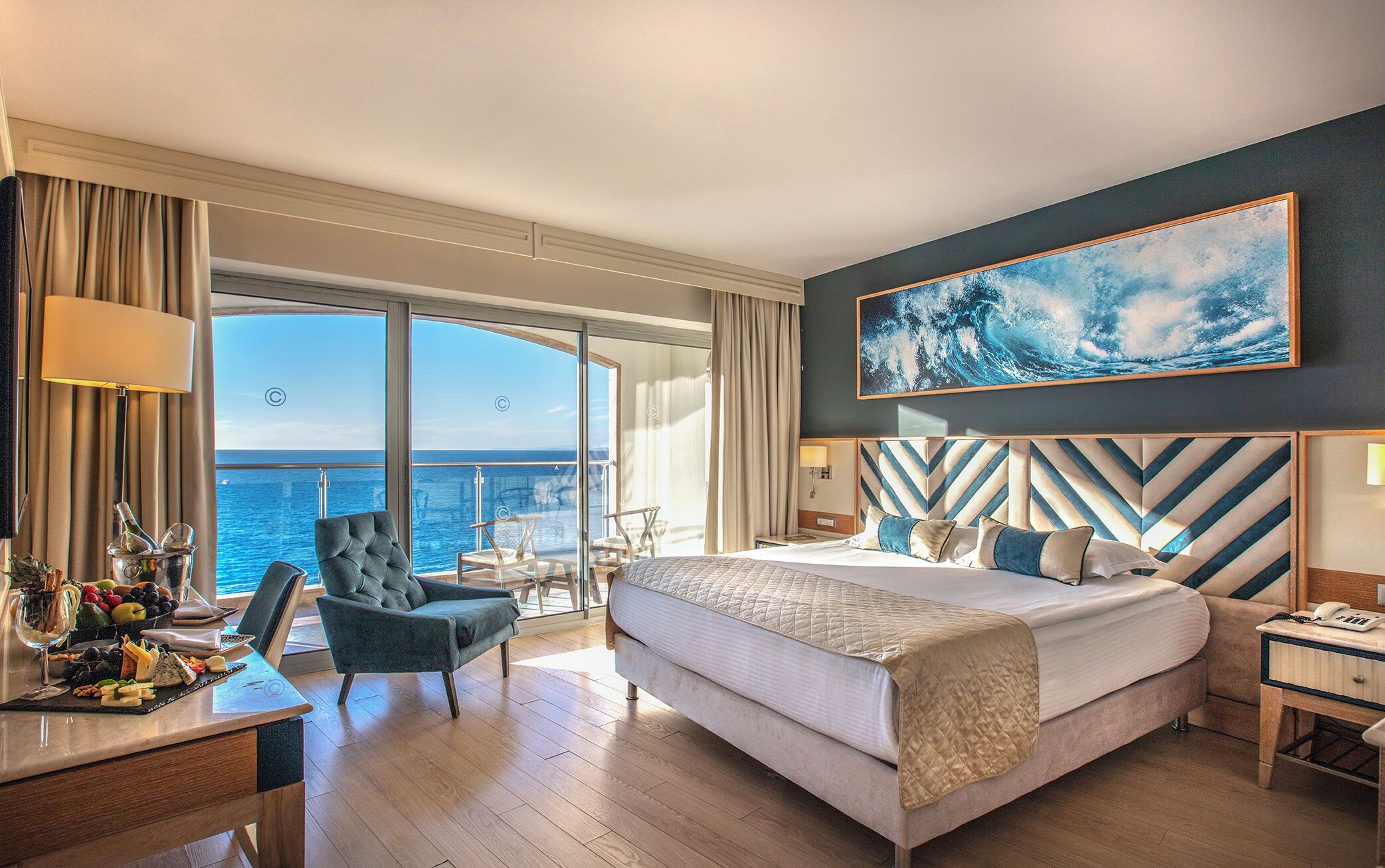 A hotel room with a large bed, a desk with a view of the sea, a chair, and a table with fruit and cheese.