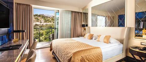 Een hotelkamer met een groot bed, een bureau met stoel, een televisie en een balkon met uitzicht op de heuvels.