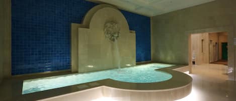 Una gran piscina interior con borde curvo, rodeada de paredes de azulejos azules y un arco de piedra.