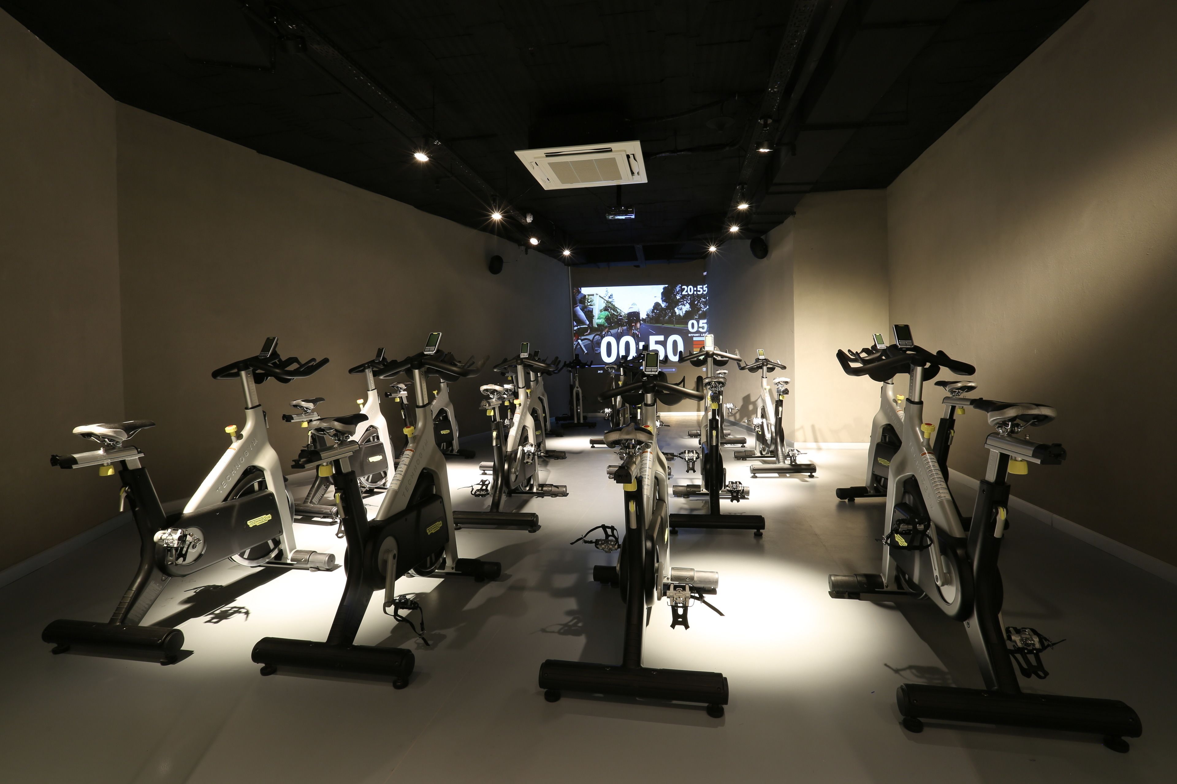 A gym with stationary bikes arranged in rows, a digital display in the background, and a ceiling with recessed lighting.