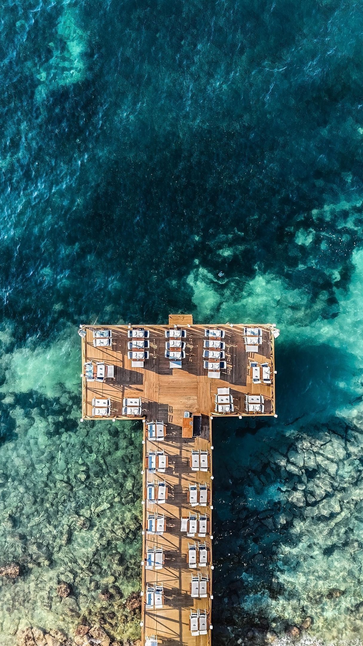 An aerial view of a T-shaped wooden deck with multiple lounge areas and a central seating area, surrounded by clear blue waters.