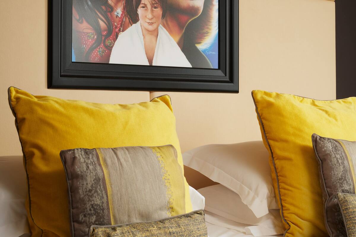 A hotel room with a bed, yellow and gray pillows, and a framed picture on the wall.