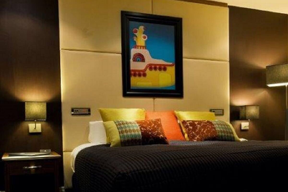 A hotel room with a large bed, multiple pillows, a nightstand, a wall-mounted lamp, and a framed picture above the bed.