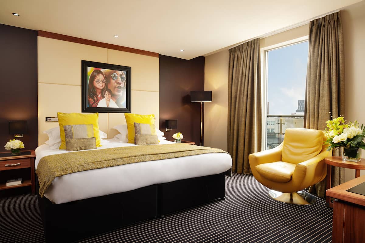 A hotel room with a large bed, a yellow armchair, a desk with a lamp, and a large window with curtains.