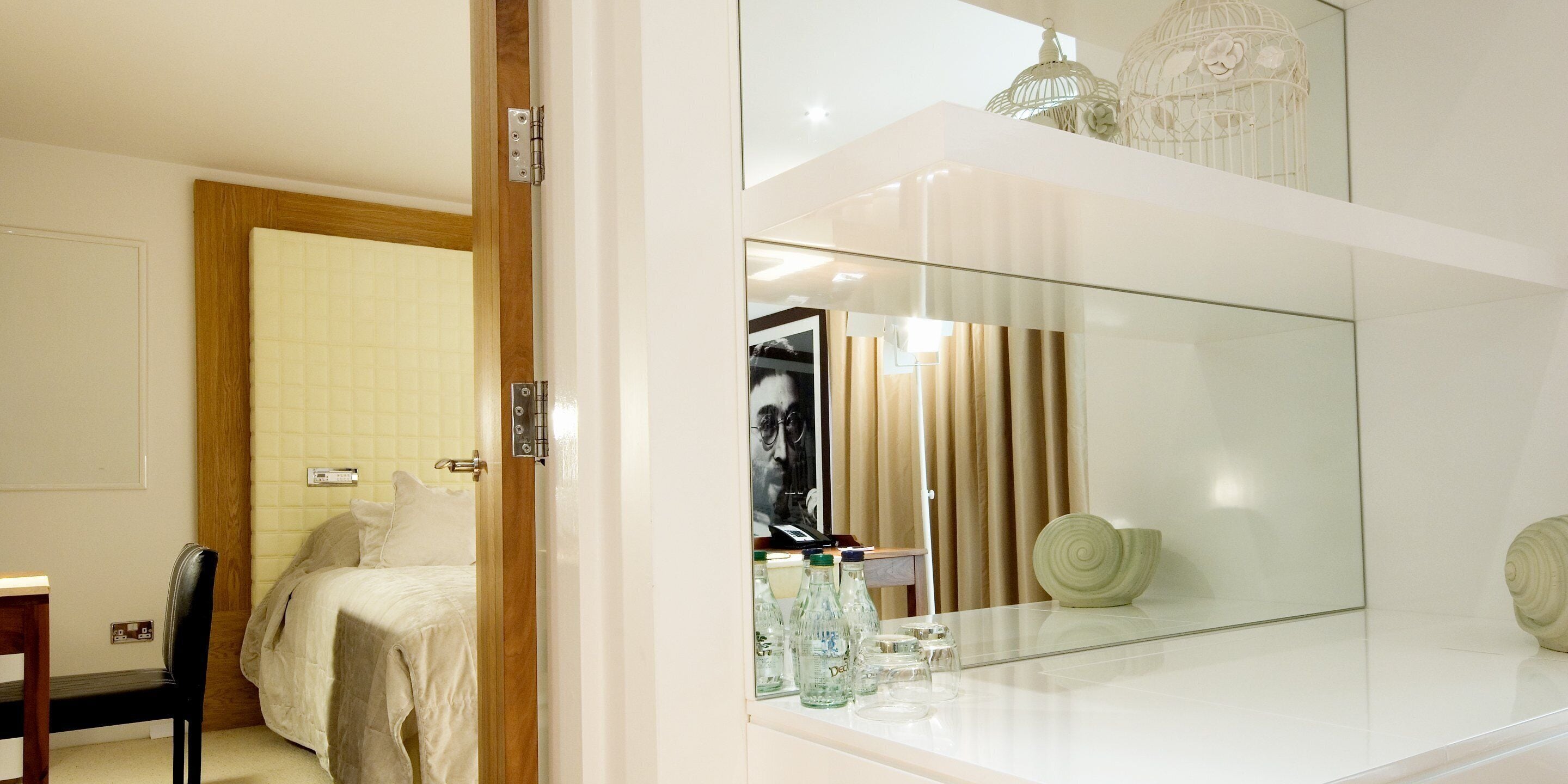 A modern bedroom with a large mirror, a bed, a chair, and a small table with bottles and glasses.